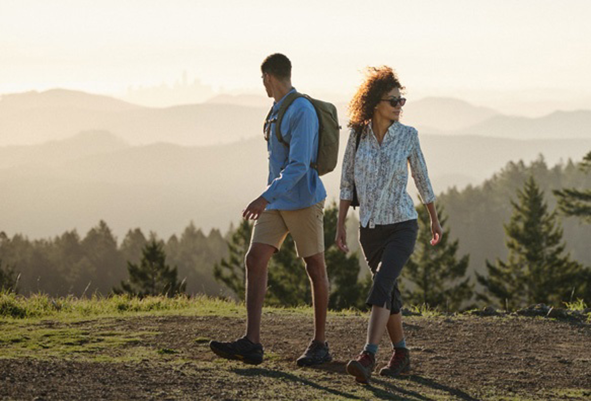 Man and woman on hiking trail overlooking hazy hills and city in background.  