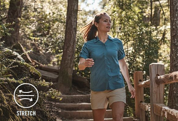 Woman in a blue button up shirt walking down stairs on a wooded path with a symbol at the bottom of the image that says stretch.
