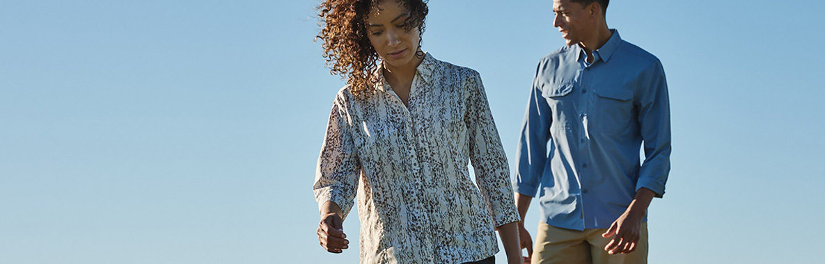 Man in blue and woman in white patterned button up shirts walking with blue sky background.