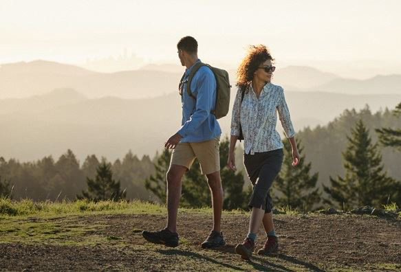 Man in blue and woman in white patterned button up shirts walking on hilltop path with evergreen trees in hazy background. 